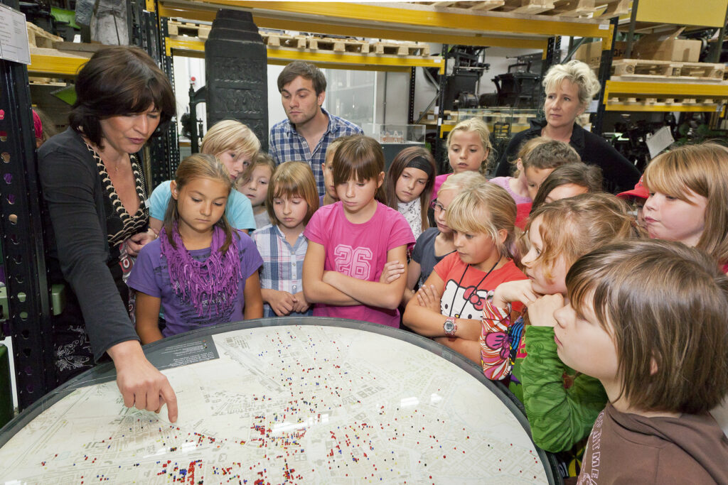 Kinder stehen um einen runden Kartentisch herum und hören den Erklärungen einer Frau zu. Im Hintergrund erkennt man Schwerlastregale.