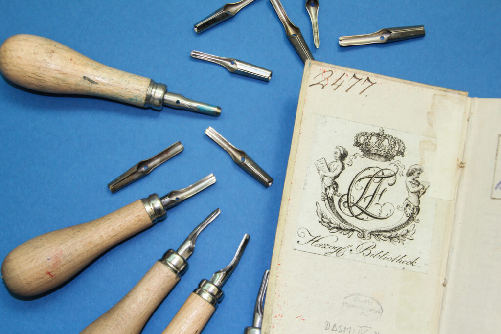 Linolschnittwerkzeuge liegen auf einer blauen Unterlage. Daneben liegt ein altes Buch mit einem kunstvoll gestaltetem Exlibris.